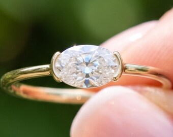 Puede incluir: Un anillo de oro con una piedra preciosa transparente de corte ovalado horizontal en un engaste de bisel. La banda es fina y pulida, mostrando un diseño sencillo que resalta las facetas y la claridad de la piedra central con elegancia.