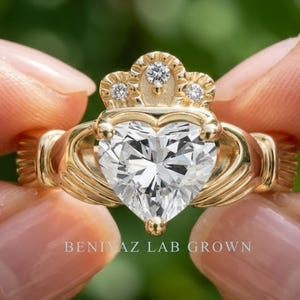 Hand held close-up of a heart cut lab grown diamond Claddagh ring showing the yellow gold crown and sculpted hands design.
