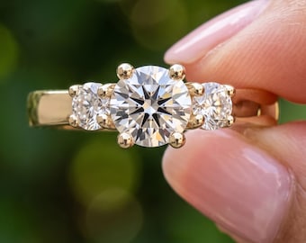 Puede incluir: Un anillo de oro con una piedra central grande de corte redondo flanqueada por dos piedras redondas más pequeñas, todas en engastes de garras. El texto "BENIYAZ LAB GROWN" está impreso en blanco en la parte inferior.