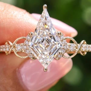 May include: A gold ring featuring a large kite-shaped center stone and smaller round accent stones. The band displays an intricate, openwork design with delicate metal loops and pavé-set diamonds extending along the sides of the ring.