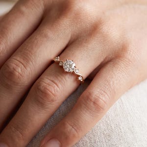 K&ouml;nnte beinhalten: Ein Ring aus Ros&eacute;gold mit einem zentralen, rund geschliffenen wei&szlig;en Edelstein, der auf jeder Seite von zwei kleineren runden wei&szlig;en Edelsteinen flankiert wird und auf einer zarten, leicht geschwungenen Ringschiene sitzt.