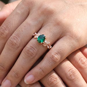 Oval lab created emerald ring worn on hand, featuring a green center stone and delicate nature inspired leaf design.