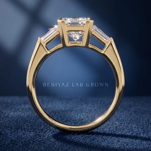Back profile of three stone emerald cut engagement ring on blue background showing open basket setting, straight shank and raised center diamond