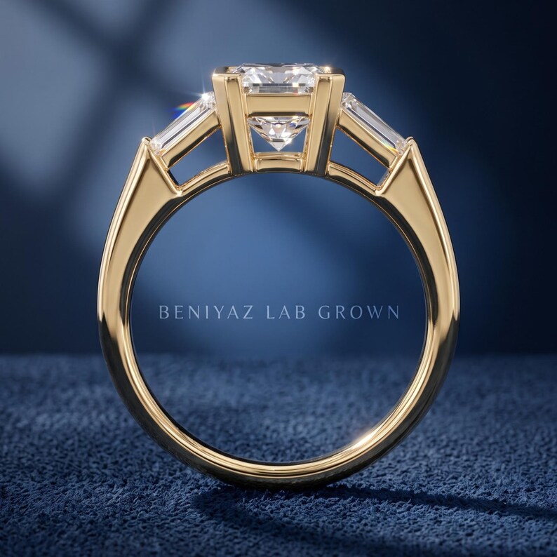 Back profile of three stone emerald cut engagement ring on blue background showing open basket setting, straight shank and raised center diamond