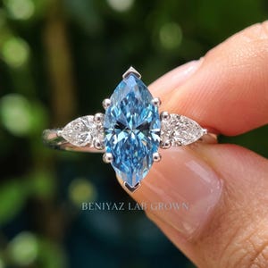 Hand held close-up of a blue marquise engagement ring with a 1.50 CT lab grown center diamond and pear shaped side stones.