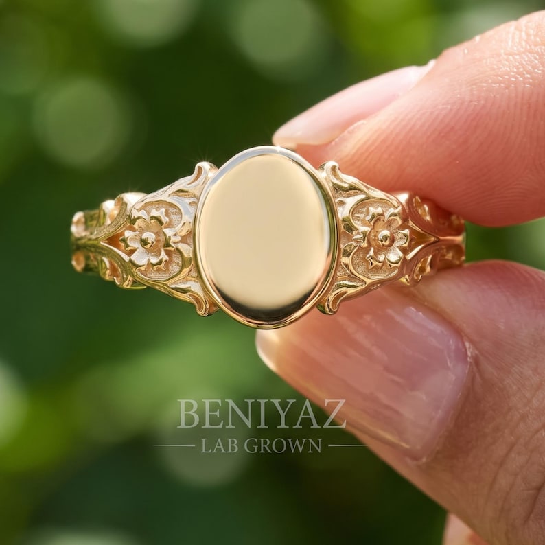 May include: A gold ring featuring a smooth, polished oval center plate flanked by intricate floral relief carvings on both sides of the band. The text BENIYAZ LAB GROWN is printed in white lettering across the lower portion of the ring surface.