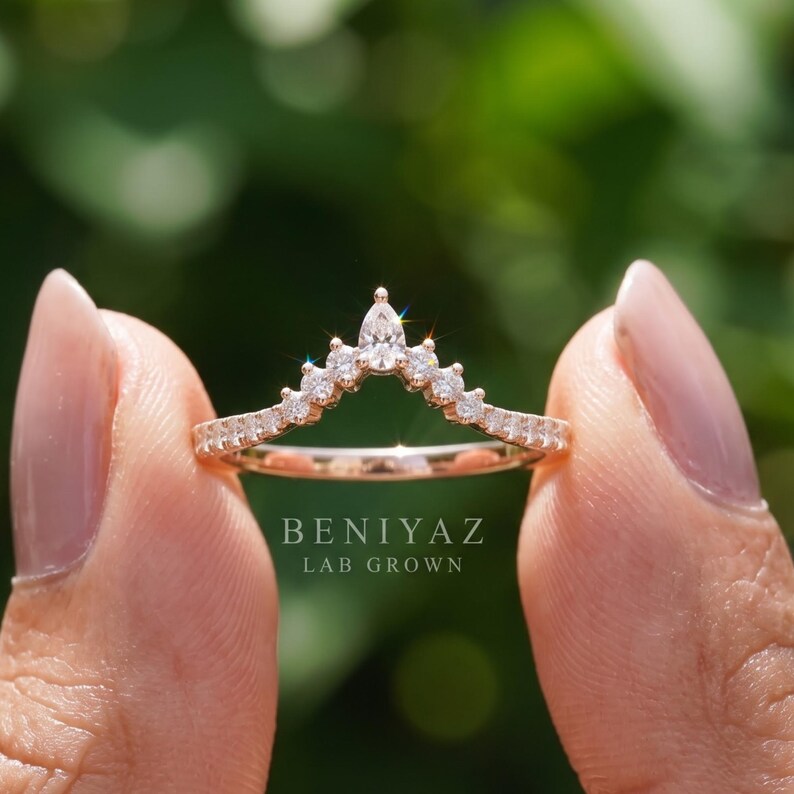 Close hand view of a 14K yellow gold contour wedding band with lab grown pear and round diamonds, elegant curved nesting ring photographed against green foliage