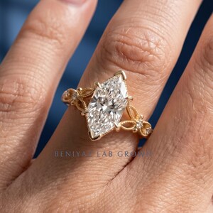 May include: A gold ring featuring a large marquise-cut center stone and intricate filigree detailing along the band, accented with small round stones. The text BENIYAZ LAB GROWN is visible at the base of the ring, showcasing a detailed metal design.