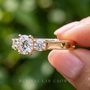 May include: A rose gold three-stone ring featuring a large round brilliant-cut centre diamond flanked by two smaller round diamonds. The band is smooth and polished. The text BENIYAZ LAB GROWN is printed at the bottom of the image.