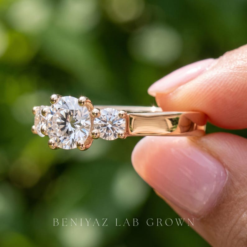 May include: A rose gold three-stone ring featuring a large round brilliant-cut centre diamond flanked by two smaller round diamonds. The band is smooth and polished. The text BENIYAZ LAB GROWN is printed at the bottom of the image.