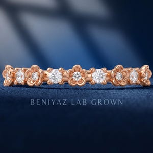 May include: A rose gold band ring featuring an alternating pattern of round clear stones and five-petaled flower motifs with a central stone. The text BENIYAZ LAB GROWN is visible below the ring, which is set with sparkling, faceted gemstones.