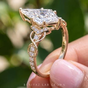 Full side view of a kite cut lab grown diamond engagement ring held upright between fingertips. The photograph displays the raised gemstone setting, decorative gallery and infinity style pavé shoulders extending down the slender gold tone shank.
