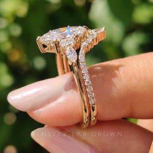 Side view of a marquise engagement ring set held between fingertips outdoors. The elevated diamond setting and two closely fitted rose gold tone bands feature sparkling accent stones along their upper sections.