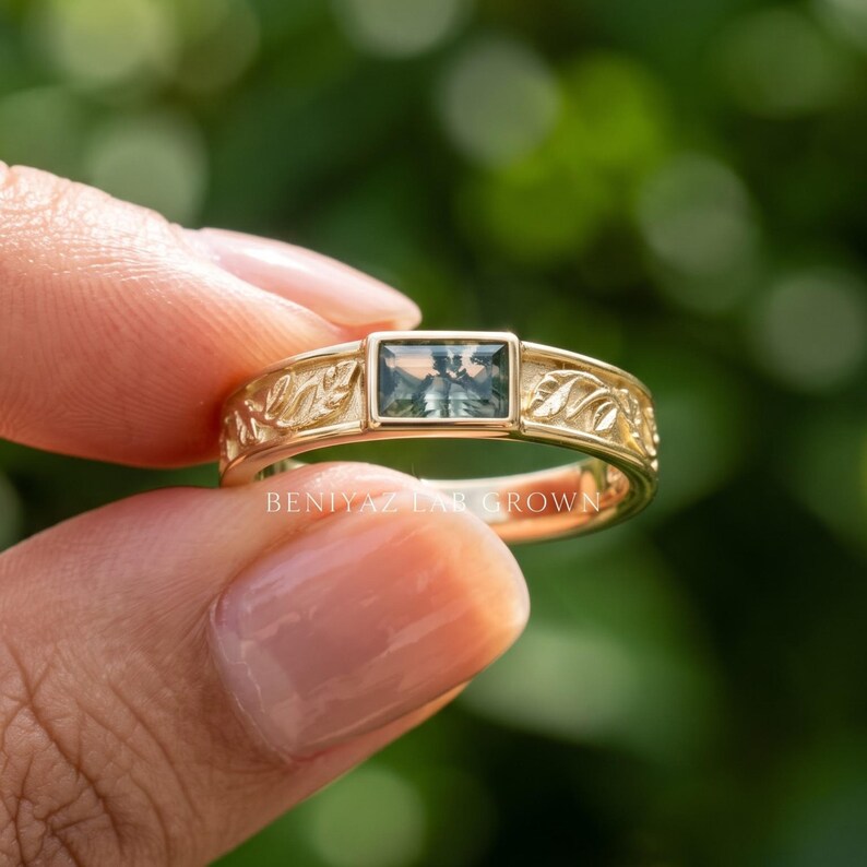 Puede incluir: Un anillo de oro con una piedra preciosa verde rectangular de corte baguette en el centro. La banda est&aacute; elaborada con un patr&oacute;n de hojas detallado y en relieve a ambos lados de la piedra. El texto "BENIYAZ LAB GROWN" es visible en la banda.