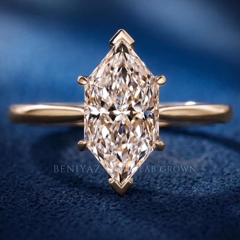 Op de afbeelding: Een ros&eacute;gouden ring met een in marquise geslepen middensteen met een gefacetteerd oppervlak, vastgehouden door zes pootjes. De ringband is glad en gepolijst. De tekst BENIYAZ LAB GROWN is zichtbaar aan de basis van de steen op het blauwe oppervlak.