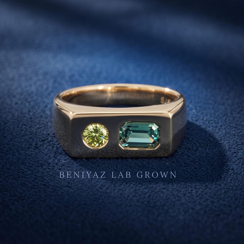 Angled product view of a two stone sapphire signet ring with teal emerald cut sapphire and round green sapphire in a polished flat top band