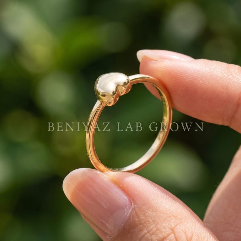 Puffy heart ring with smooth polished heart centerpiece and slim band, a dainty minimalist love ring held between fingertips.