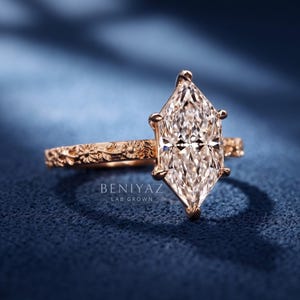 Close up hand held marquise engagement ring with an elongated lab grown diamond center and decorative floral band.