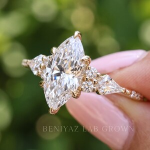 Angled front photograph of a marquise diamond engagement ring against a softly blurred garden background. The bright center diamond, geometric accent stones and detailed gold tone shoulders are shown in close-up.