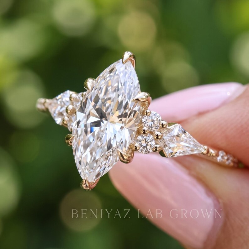 Angled front photograph of a marquise diamond engagement ring against a softly blurred garden background. The bright center diamond, geometric accent stones and detailed gold tone shoulders are shown in close-up.