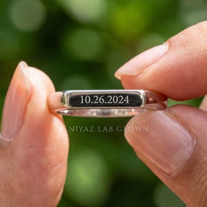 Hand displaying a custom engraved band with personalized text detail, modern minimalist wedding ring shown against soft green foliage