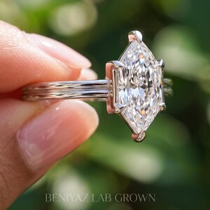 Angled macro photograph of an elongated Dutch marquise lab grown diamond ring, showing the sparkling faceted center diamond, six-prong solitaire mounting and sleek double-line band. Elegant minimalist engagement ring held gently between fingers.