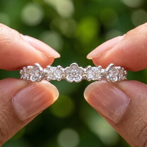 May include: A silver-toned band ring featuring a repeating pattern of small, five-petaled flower shapes and round, clear, faceted stones. The metal band is polished and reflective, with each flower setting containing a central sparkling stone.
