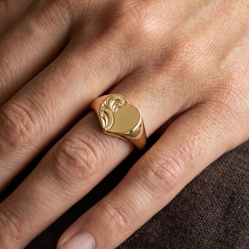 Close up of a heart signet pinky ring worn on hand, featuring a polished heart face and delicate vintage floral engraving