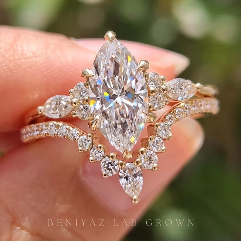 Front-facing macro photograph of a vintage inspired marquise diamond bridal set held between fingertips. A brilliant marquise center stone sits above an ornate diamond-accented engagement ring, complemented by a curved matching wedding band with sparkling stones.