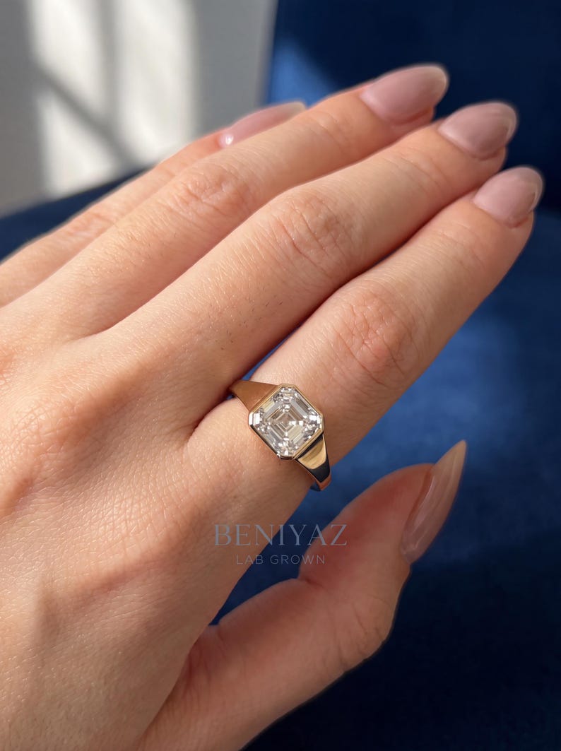 Top-view hand photo featuring a 1.55 CT Asscher cut lab grown diamond ring in 14K yellow gold. The bezel set signet engagement ring is worn on the ring finger and displays an IGI certified D color VVS1 lab grown diamond, combining a modern statement ring look with a clean luxury aesthetic.