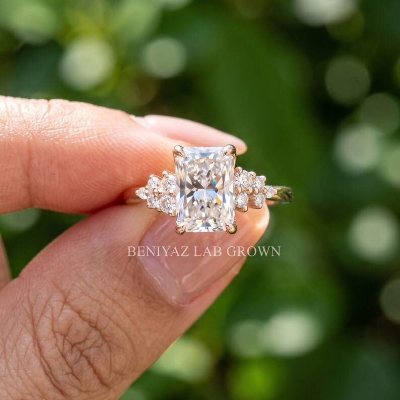 May include: A radiant-cut center stone ring set in a gold band with smaller round accent stones on each side. The ring features the text BENIYAZ LAB GROWN engraved on the band. The design includes a cluster of three small stones flanking the center.