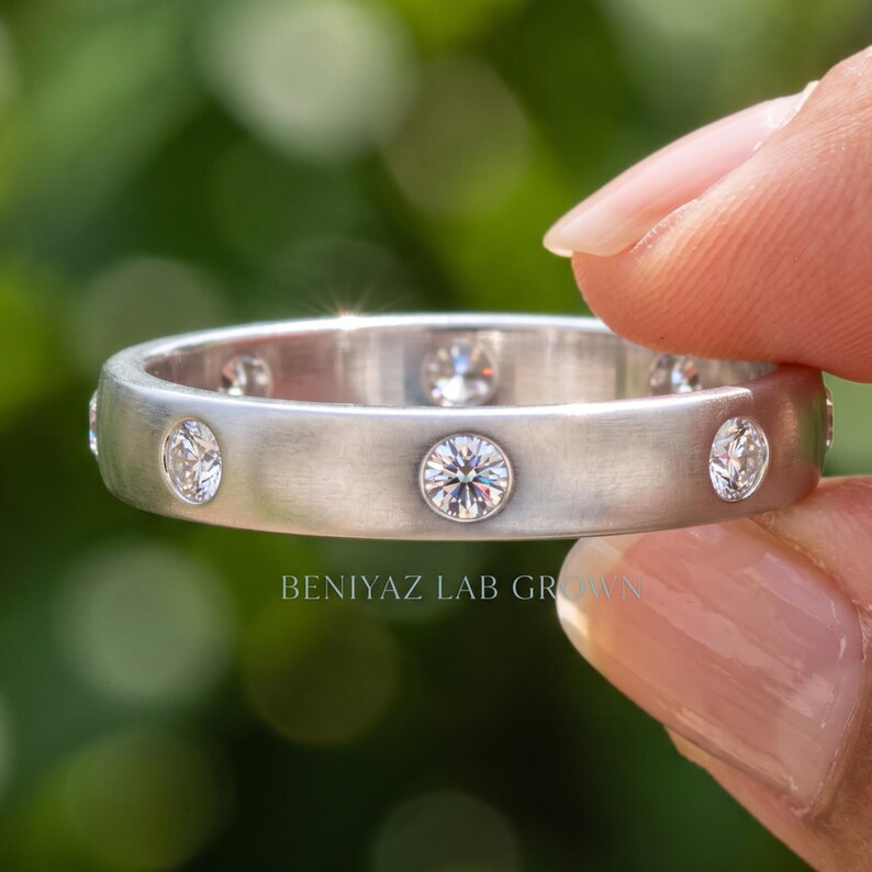 Hand displaying a lab grown diamond wedding band in polished white gold, featuring evenly spaced round bezel set diamonds against a soft green background