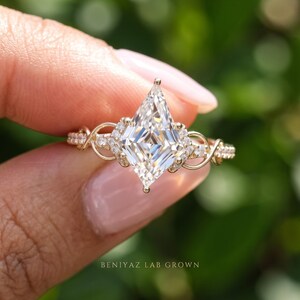 Three-quarter front view of a kite shaped lab grown diamond ring held delicately between fingers. The photograph highlights the distinctive angular center stone, brilliant diamond accents and intertwined pavé detailing along the gold tone band in soft outdoor light.