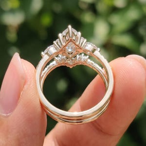 Upright rear view of a marquise engagement ring and matching curved wedding band held between fingertips. The open setting beneath the center diamond, decorative gallery and circular ring shanks are visible against blurred greenery.