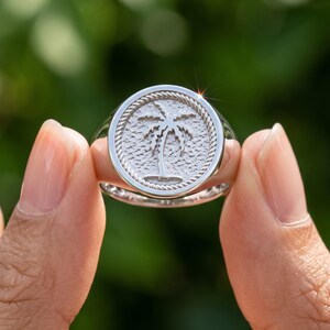 Hand displaying a sterling silver palm tree signet ring with circular medallion face, tropical palm motif and decorative rope border