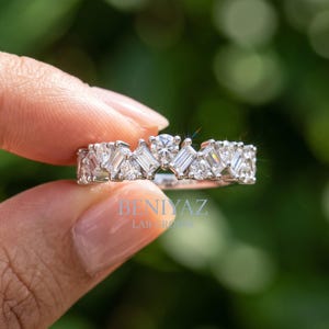 May include: A silver-toned ring featuring a band set with alternating round and baguette-cut clear stones. The text BENIYAZ LAB GROWN is printed in light blue on the center of the band. The design showcases a repeating pattern of faceted gems.