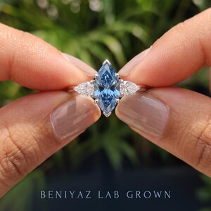Hand held side view of a three stone blue marquise diamond engagement ring showing the slim band and raised center setting.