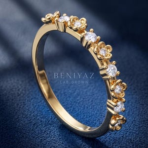 May include: A gold ring featuring a series of alternating flower-shaped settings and round prong-set stones. The band is polished gold, and the text BENIYAZ LAB GROWN is visible in the center. The design incorporates multiple clear, sparkling stones.