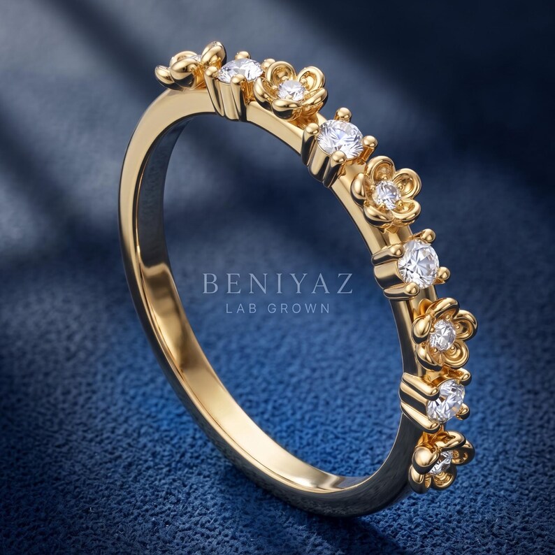May include: A gold ring featuring a series of alternating flower-shaped settings and round prong-set stones. The band is polished gold, and the text BENIYAZ LAB GROWN is visible in the center. The design incorporates multiple clear, sparkling stones.