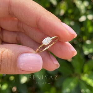 Close-up of a hand holding a minimalist oval bezel diamond ring against a naturally blurred green background. The polished gold band and horizontally set oval gemstone are visible alongside soft pink manicured fingernails.