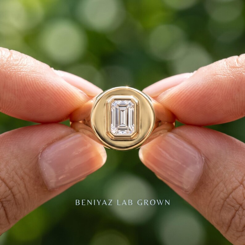 Hand held yellow gold emerald cut lab grown diamond signet ring showing the center stone and wide signet shoulders.