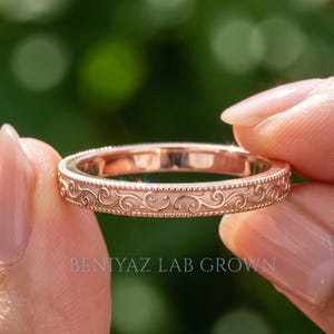 May include: A rose gold band ring featuring an intricate engraved scrollwork pattern along the outer surface, bordered by delicate beaded edges. The text BENIYAZ LAB GROWN is printed in light gray at the bottom of the image.