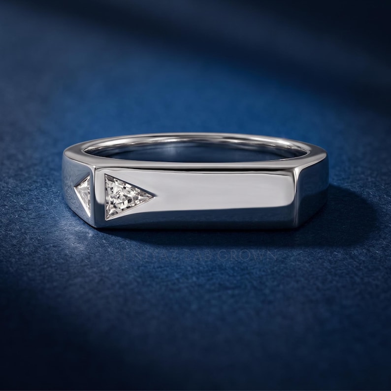 Puede incluir: Un anillo de metal plateado con una superficie superior plana y rectangular, con dos peque&ntilde;as piedras transparentes de corte triangular incrustadas en el lateral. La banda tiene un acabado pulido y liso con un dise&ntilde;o geom&eacute;trico de bordes definidos.