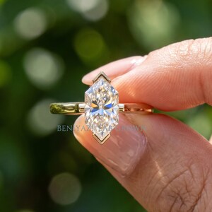 May include: A gold ring featuring a marquise-cut center stone held in a bezel setting with pointed ends. The band is smooth and polished gold. The text BENIYAZI LAB GROWN is visible on the ring, which displays a clear, faceted gemstone.