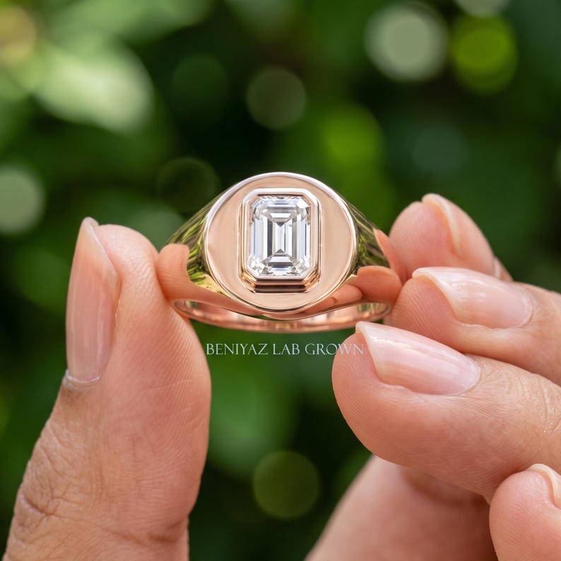 Hand held rose gold emerald cut diamond signet ring showing the vertical rectangular center stone and polished face.