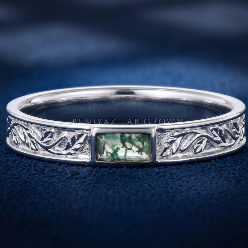 Puede incluir: Un anillo de tono plateado con una piedra preciosa rectangular de &aacute;gata musgosa en el centro. La banda est&aacute; decorada con un patr&oacute;n de hojas en relieve a ambos lados de la piedra. El texto BENIYAZ LAB GROWN es visible en la superficie interior.