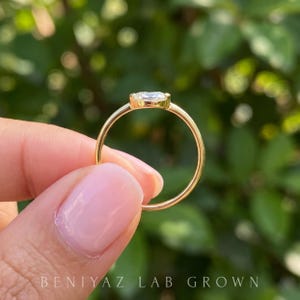 Three-quarter view of an east west oval lab grown diamond ring held delicately between fingertips. The horizontal oval center stone, smooth gold bezel and thin polished shank are shown in natural daylight with greenery behind.