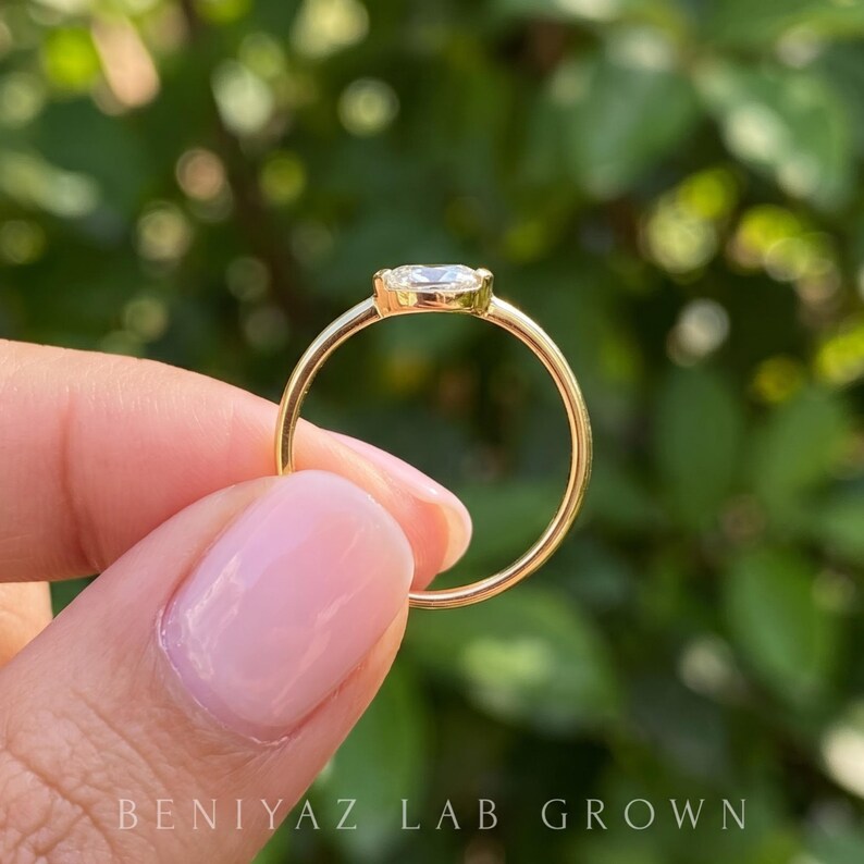 Three-quarter view of an east west oval lab grown diamond ring held delicately between fingertips. The horizontal oval center stone, smooth gold bezel and thin polished shank are shown in natural daylight with greenery behind.