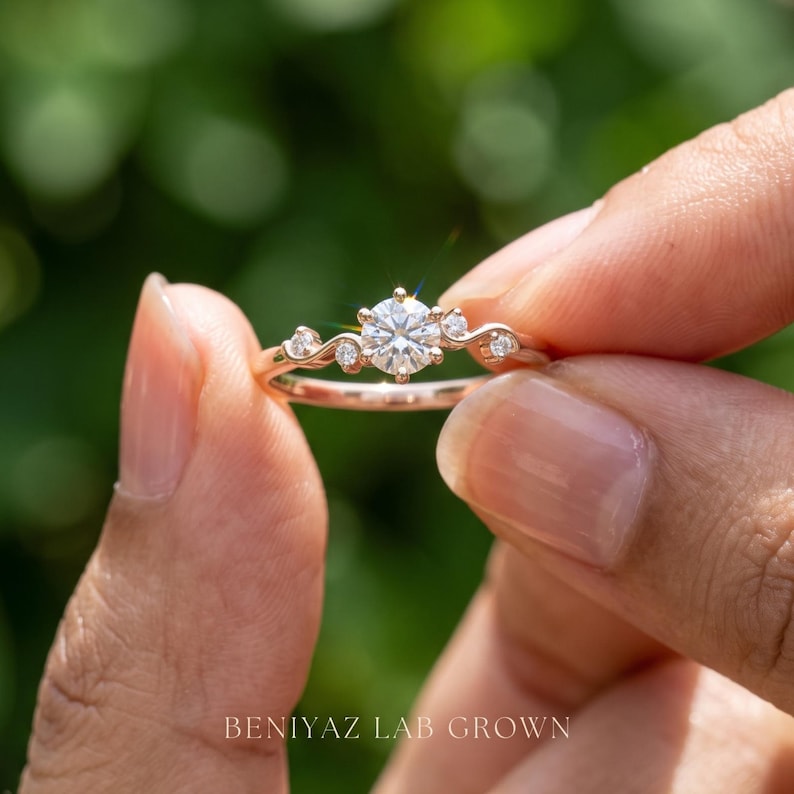 K&ouml;nnte beinhalten: Ein Ring aus Ros&eacute;gold mit einem zentralen runden Brillantschliff-Stein, flankiert von zwei kleineren Steinen auf jeder Seite, eingefasst in eine zarte, geschwungene Ringschiene. Der Text BENIYAZ LAB GROWN ist unten zu sehen.