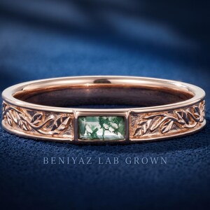 Puede incluir: Un anillo de oro rosa con una piedra de &aacute;gata musgosa rectangular en el centro, flanqueada por patrones de hojas grabados a lo largo de la banda. El texto BENIYAZ LAB GROWN aparece debajo del anillo, que tiene un acabado met&aacute;lico pulido.